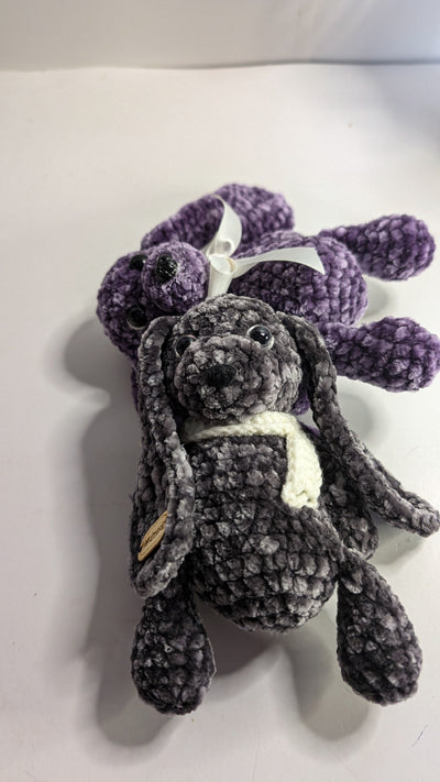 Two crochet bunnies from FashionableCrochet—one gray with a white scarf, the other purple with a white bow—rest on a white surface. These handmade toys boast long ears and round bodies, meticulously crafted from soft yarn, making them delightful and decorative pieces for any space.