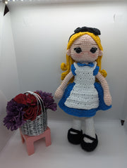 The Crochet Alice Amigurumi Doll by FashionableCrochet features a handmade design with yellow hair, a black bow, and a blue dress complemented by a white apron, reminiscent of an Alice in Wonderland collectible. She stands beside a small pink stool holding a basket of red and purple flowers.