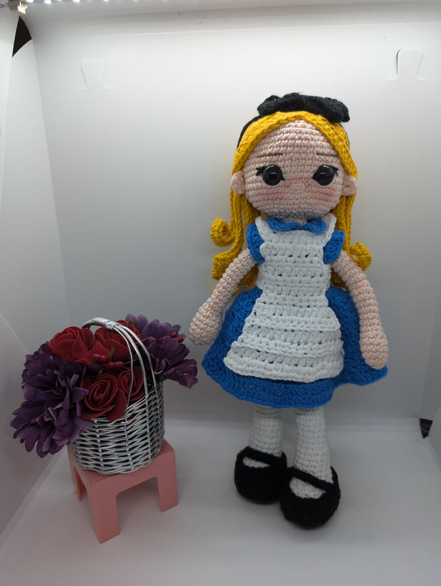 The Crochet Alice Amigurumi Doll by FashionableCrochet features a handmade design with yellow hair, a black bow, and a blue dress complemented by a white apron, reminiscent of an Alice in Wonderland collectible. She stands beside a small pink stool holding a basket of red and purple flowers.