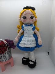 The Crochet Alice Amigurumi Doll by FashionableCrochet features a handmade design with yellow hair, a black bow, and a blue dress complemented by a white apron, reminiscent of an Alice in Wonderland collectible. She stands beside a small pink stool holding a basket of red and purple flowers.