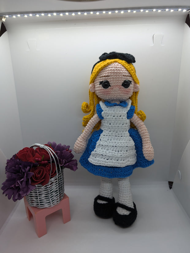 The Crochet Alice Amigurumi Doll by FashionableCrochet features a handmade design with yellow hair, a black bow, and a blue dress complemented by a white apron, reminiscent of an Alice in Wonderland collectible. She stands beside a small pink stool holding a basket of red and purple flowers.