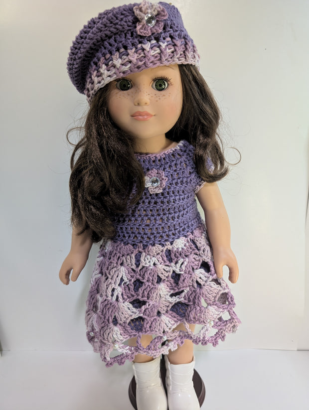 An elegantly displayed amigurumi doll from FashionableCrochet showcases the art of handmade crochet doll clothes, featuring a purple crocheted dress and matching beret adorned with floral accents. Her long, dark hair and white boots complete her stylish look.