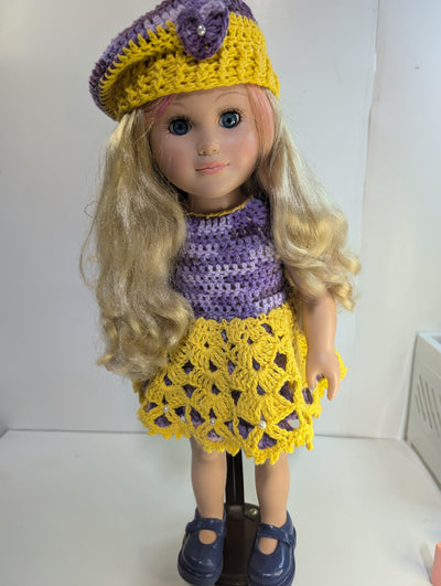 A doll with long blonde hair is dressed in FashionableCrochet's Handmade Crochet Doll Clothes, featuring a purple and yellow dress with a matching beret embellished with a flower. It is displayed wearing dark shoes against a plain background, highlighting the intricate artistry of amigurumi dolls.