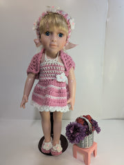 A delightful amigurumi doll with blonde hair is dressed in FashionableCrochet's handmade crochet outfit, featuring a pink and white dress, matching headband, sweater, and slippers. It stands on a wooden base next to a small pink stool adorned with a basket containing purple flowers and red apples.