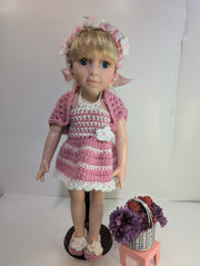 A delightful amigurumi doll with blonde hair is dressed in FashionableCrochet's handmade crochet outfit, featuring a pink and white dress, matching headband, sweater, and slippers. It stands on a wooden base next to a small pink stool adorned with a basket containing purple flowers and red apples.