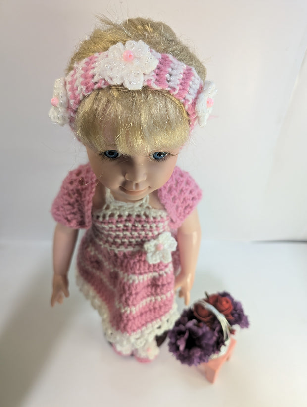 A delightful amigurumi doll with blonde hair is dressed in FashionableCrochet's handmade crochet outfit, featuring a pink and white dress, matching headband, sweater, and slippers. It stands on a wooden base next to a small pink stool adorned with a basket containing purple flowers and red apples.