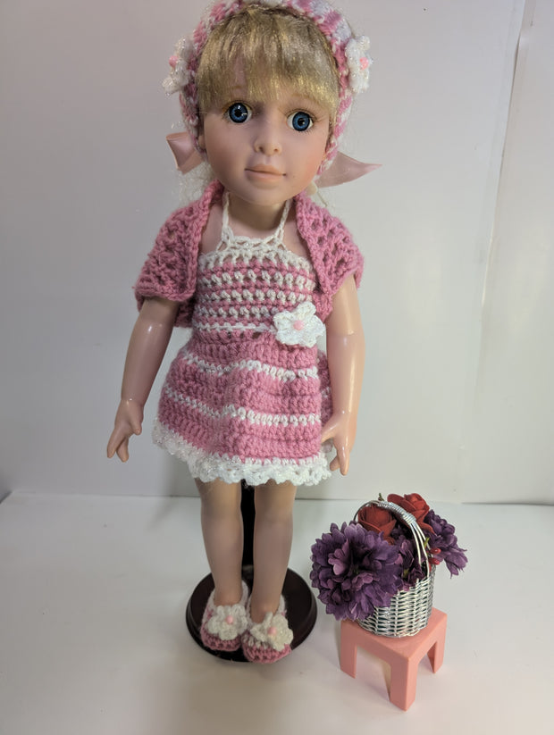 A delightful amigurumi doll with blonde hair is dressed in FashionableCrochet's handmade crochet outfit, featuring a pink and white dress, matching headband, sweater, and slippers. It stands on a wooden base next to a small pink stool adorned with a basket containing purple flowers and red apples.