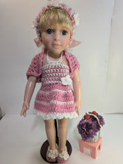 A delightful amigurumi doll with blonde hair is dressed in FashionableCrochet's handmade crochet outfit, featuring a pink and white dress, matching headband, sweater, and slippers. It stands on a wooden base next to a small pink stool adorned with a basket containing purple flowers and red apples.