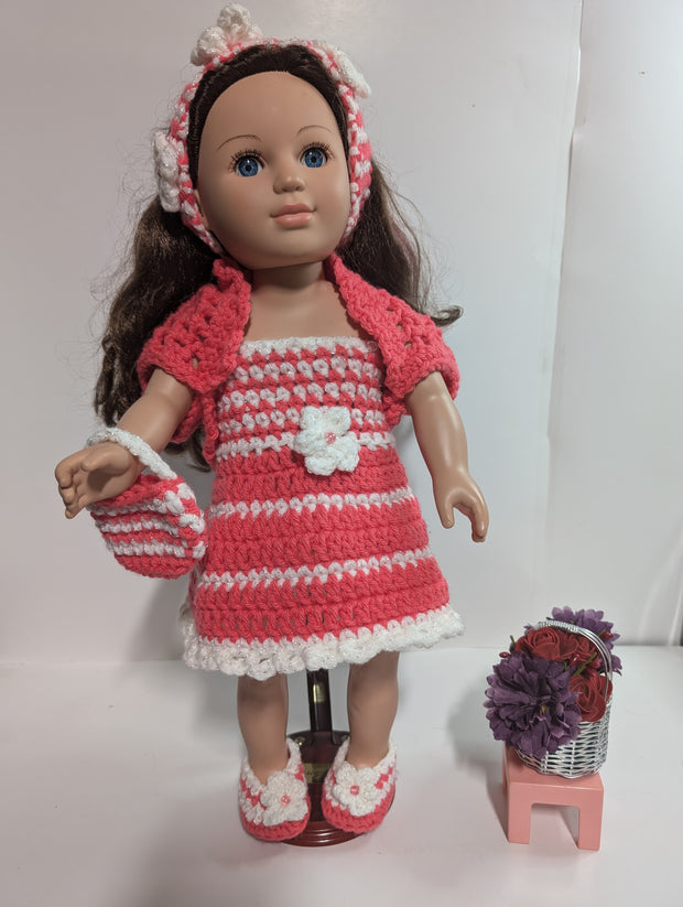 The FashionableCrochet Handmade Crochet Doll Clothes set features a doll adorned with long brown hair, dressed in a delightful pink and white striped dress, cardigan, hairband, and shoes. The ensemble is completed with a coordinating handbag. Accompanying the doll is a small stool that supports a basket brimming with purple and red flowers.