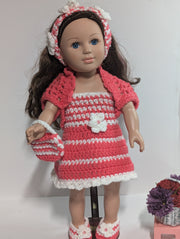 The FashionableCrochet Handmade Crochet Doll Clothes set features a doll adorned with long brown hair, dressed in a delightful pink and white striped dress, cardigan, hairband, and shoes. The ensemble is completed with a coordinating handbag. Accompanying the doll is a small stool that supports a basket brimming with purple and red flowers.