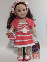 The FashionableCrochet Handmade Crochet Doll Clothes set features a doll adorned with long brown hair, dressed in a delightful pink and white striped dress, cardigan, hairband, and shoes. The ensemble is completed with a coordinating handbag. Accompanying the doll is a small stool that supports a basket brimming with purple and red flowers.