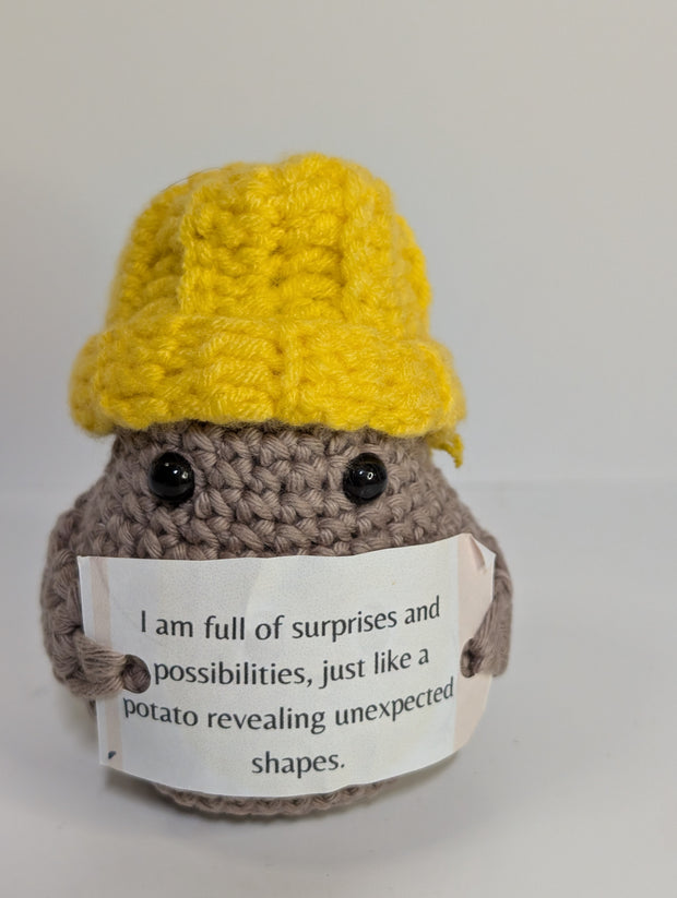 A FashionableCrochet Handmade Crochet Potato with black eyes holds a sign that reads, "Positive potato: I may be tiny, but I believe in you. Go do your thing." Wearing a charming white hat with a red top, this quirky amigurumi toy makes for a delightful personalized gift against a plain white background.