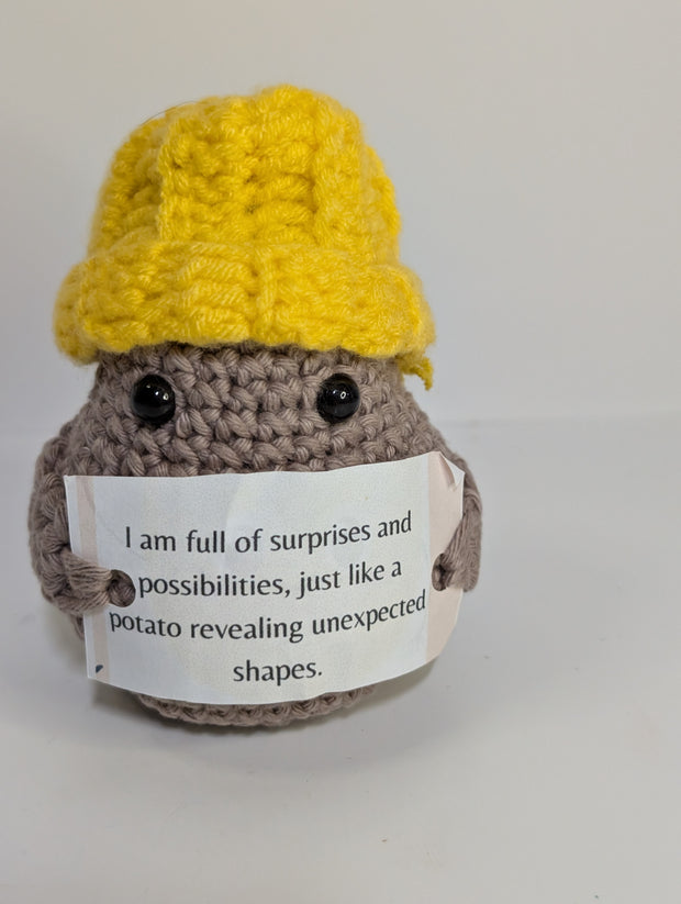 A FashionableCrochet Handmade Crochet Potato with black eyes holds a sign that reads, "Positive potato: I may be tiny, but I believe in you. Go do your thing." Wearing a charming white hat with a red top, this quirky amigurumi toy makes for a delightful personalized gift against a plain white background.