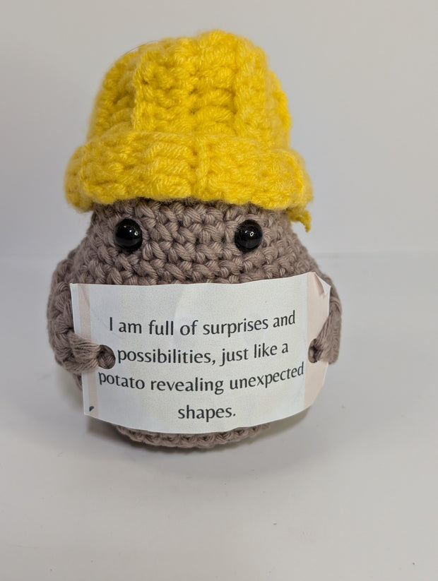 A FashionableCrochet Handmade Crochet Potato with black eyes holds a sign that reads, "Positive potato: I may be tiny, but I believe in you. Go do your thing." Wearing a charming white hat with a red top, this quirky amigurumi toy makes for a delightful personalized gift against a plain white background.