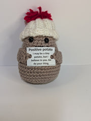 A FashionableCrochet Handmade Crochet Potato with black eyes holds a sign that reads, "Positive potato: I may be tiny, but I believe in you. Go do your thing." Wearing a charming white hat with a red top, this quirky amigurumi toy makes for a delightful personalized gift against a plain white background.