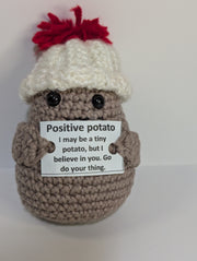 A FashionableCrochet Handmade Crochet Potato with black eyes holds a sign that reads, "Positive potato: I may be tiny, but I believe in you. Go do your thing." Wearing a charming white hat with a red top, this quirky amigurumi toy makes for a delightful personalized gift against a plain white background.