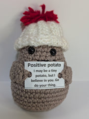 A FashionableCrochet Handmade Crochet Potato with black eyes holds a sign that reads, "Positive potato: I may be tiny, but I believe in you. Go do your thing." Wearing a charming white hat with a red top, this quirky amigurumi toy makes for a delightful personalized gift against a plain white background.