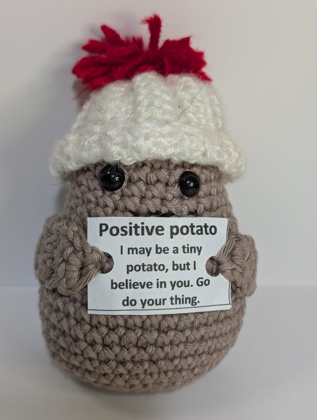 A FashionableCrochet Handmade Crochet Potato with black eyes holds a sign that reads, "Positive potato: I may be tiny, but I believe in you. Go do your thing." Wearing a charming white hat with a red top, this quirky amigurumi toy makes for a delightful personalized gift against a plain white background.