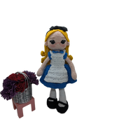 The Crochet Alice Amigurumi Doll by FashionableCrochet features a handmade design with yellow hair, a black bow, and a blue dress complemented by a white apron, reminiscent of an Alice in Wonderland collectible. She stands beside a small pink stool holding a basket of red and purple flowers.