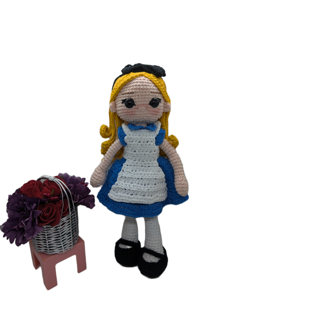 The Crochet Alice Amigurumi Doll by FashionableCrochet features a handmade design with yellow hair, a black bow, and a blue dress complemented by a white apron, reminiscent of an Alice in Wonderland collectible. She stands beside a small pink stool holding a basket of red and purple flowers.